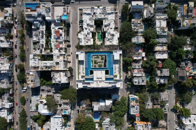Aerial view of the fully constructed Ceiba at 25 luxury residential building in Playa del Carmen, showcasing its modern white facade, multiple levels of balconies with glass railings, and a prominent rooftop featuring a large, blue-tiled swimming ...