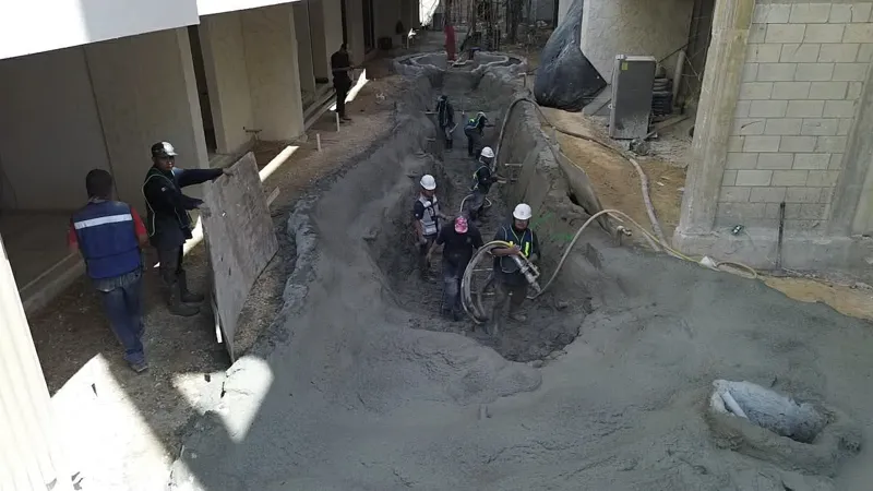 Construction workers in hard hats and safety vests are actively pouring and shaping wet concrete into a long, winding trench, forming a water feature or lazy river at the Zamajal development. Rebar is visible for structural support, with surroundi...