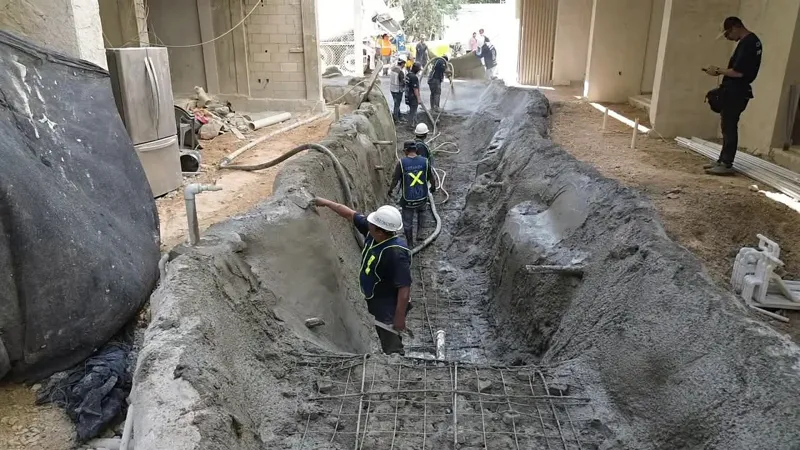 Construction workers in hard hats and safety vests are actively pouring and shaping wet concrete into a long, winding trench, forming a water feature or lazy river at the Zamajal development. Rebar is visible for structural support, with surroundi...