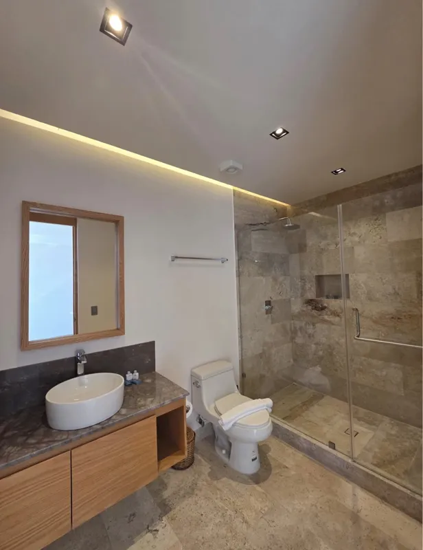 A bright, modern bathroom in an Akua Signature model unit, featuring a floating light wood vanity with a white vessel sink, a large rectangular mirror, a white toilet with a small towel animal, and a spacious walk-in shower with a glass enclosure ...