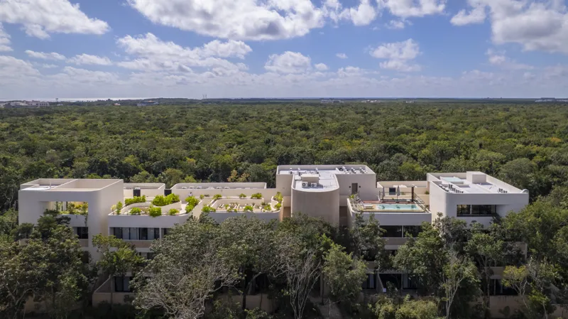 Aerial view of the Peregrina development in Tulum, showing multiple completed buildings with white facades, rooftop gardens, and a visible rooftop pool, surrounded by dense green jungle foliage under a blue sky with white clouds. The ocean is visi...