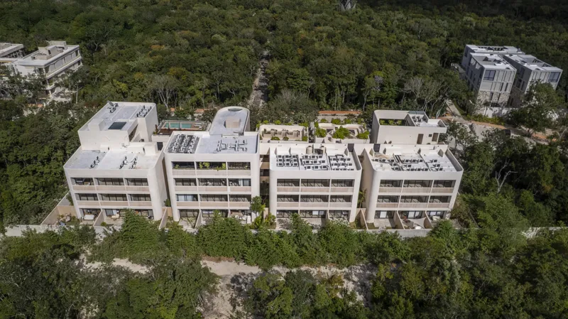 Aerial view of the Peregrina development in Tulum, showing multiple completed buildings with white facades, rooftop gardens, and a visible rooftop pool, surrounded by dense green jungle foliage under a blue sky with white clouds. The ocean is visi...