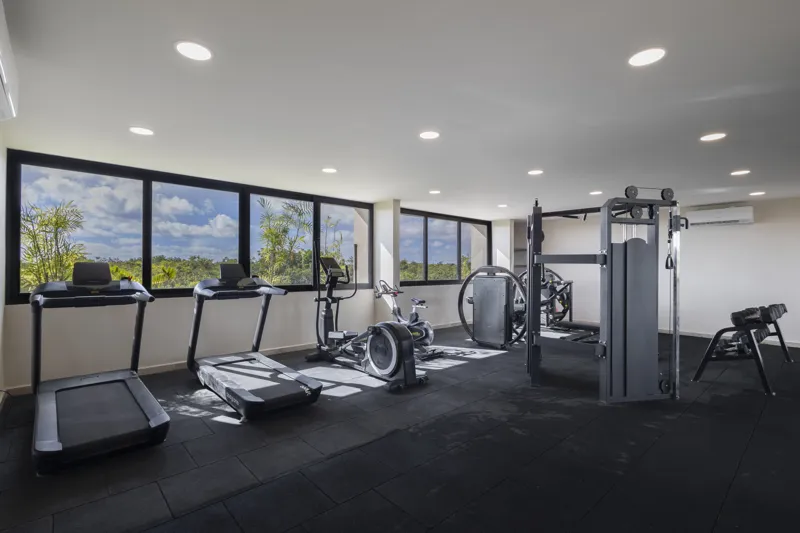 Aerial view of the Peregrina development in Tulum, showing multiple completed buildings with white facades, rooftop gardens, and a visible rooftop pool, surrounded by dense green jungle foliage under a blue sky with white clouds. The ocean is visi...