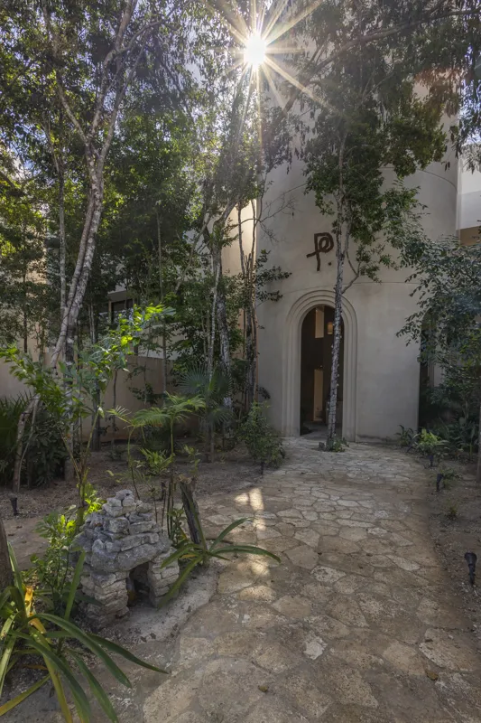 Aerial view of the Peregrina development in Tulum, showing multiple completed buildings with white facades, rooftop gardens, and a visible rooftop pool, surrounded by dense green jungle foliage under a blue sky with white clouds. The ocean is visi...