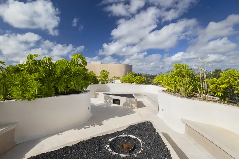 Aerial view of the Peregrina development in Tulum, showing multiple completed buildings with white facades, rooftop gardens, and a visible rooftop pool, surrounded by dense green jungle foliage under a blue sky with white clouds. The ocean is visi...