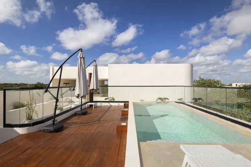 Aerial view of the Peregrina development in Tulum, showing multiple completed buildings with white facades, rooftop gardens, and a visible rooftop pool, surrounded by dense green jungle foliage under a blue sky with white clouds. The ocean is visi...