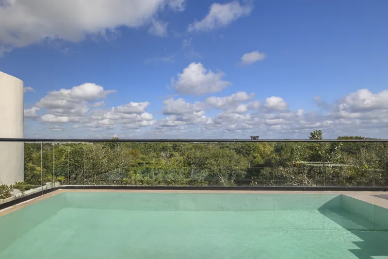 Aerial view of the Peregrina development in Tulum, showing multiple completed buildings with white facades, rooftop gardens, and a visible rooftop pool, surrounded by dense green jungle foliage under a blue sky with white clouds. The ocean is visi...