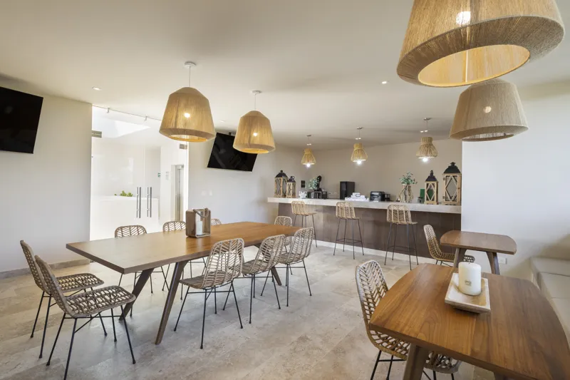 Aerial view of the Peregrina development in Tulum, showing multiple completed buildings with white facades, rooftop gardens, and a visible rooftop pool, surrounded by dense green jungle foliage under a blue sky with white clouds. The ocean is visi...