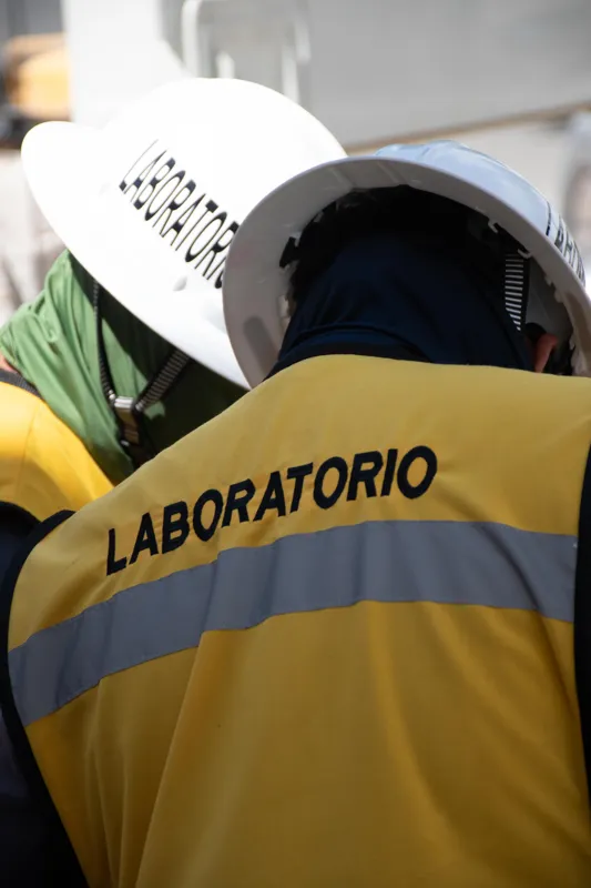 An elevated view of the Abund construction site in Riviera Maya, showing a large white 'Parma' concrete pump truck with its boom extended, actively pouring concrete into a vast, newly formed structural slab. Numerous construction workers in safety...