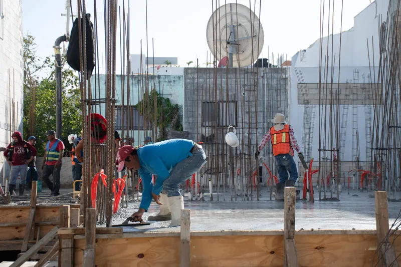 An elevated view of the Abund construction site in Riviera Maya, showing a large white 'Parma' concrete pump truck with its boom extended, actively pouring concrete into a vast, newly formed structural slab. Numerous construction workers in safety...