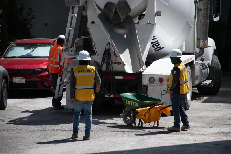 An elevated view of the Abund construction site in Riviera Maya, showing a large white 'Parma' concrete pump truck with its boom extended, actively pouring concrete into a vast, newly formed structural slab. Numerous construction workers in safety...