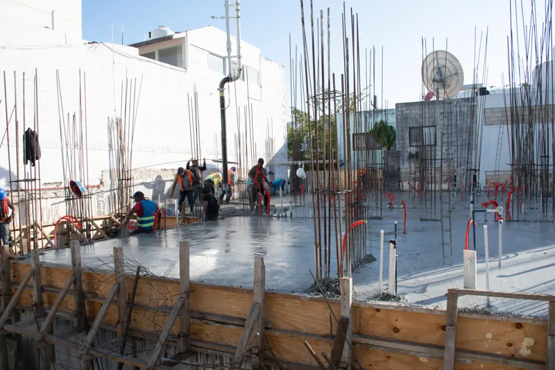 An elevated view of the Abund construction site in Riviera Maya, showing a large white 'Parma' concrete pump truck with its boom extended, actively pouring concrete into a vast, newly formed structural slab. Numerous construction workers in safety...
