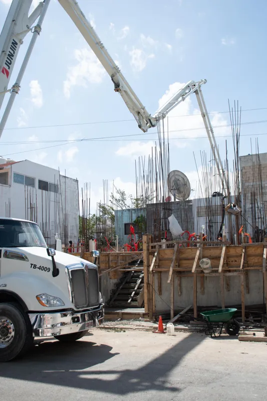 An elevated view of the Abund construction site in Riviera Maya, showing a large white 'Parma' concrete pump truck with its boom extended, actively pouring concrete into a vast, newly formed structural slab. Numerous construction workers in safety...