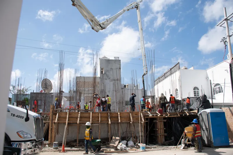 An elevated view of the Abund construction site in Riviera Maya, showing a large white 'Parma' concrete pump truck with its boom extended, actively pouring concrete into a vast, newly formed structural slab. Numerous construction workers in safety...