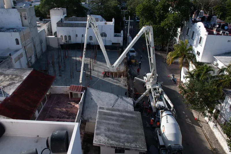 An elevated view of the Abund construction site in Riviera Maya, showing a large white 'Parma' concrete pump truck with its boom extended, actively pouring concrete into a vast, newly formed structural slab. Numerous construction workers in safety...