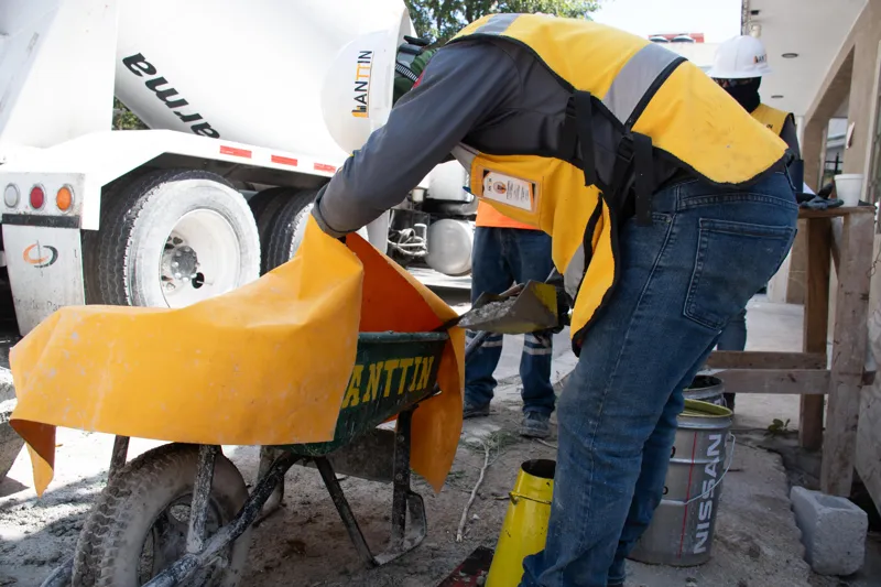 An elevated view of the Abund construction site in Riviera Maya, showing a large white 'Parma' concrete pump truck with its boom extended, actively pouring concrete into a vast, newly formed structural slab. Numerous construction workers in safety...