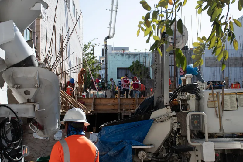 An elevated view of the Abund construction site in Riviera Maya, showing a large white 'Parma' concrete pump truck with its boom extended, actively pouring concrete into a vast, newly formed structural slab. Numerous construction workers in safety...