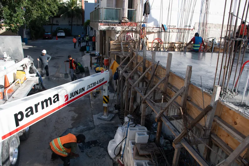 An elevated view of the Abund construction site in Riviera Maya, showing a large white 'Parma' concrete pump truck with its boom extended, actively pouring concrete into a vast, newly formed structural slab. Numerous construction workers in safety...