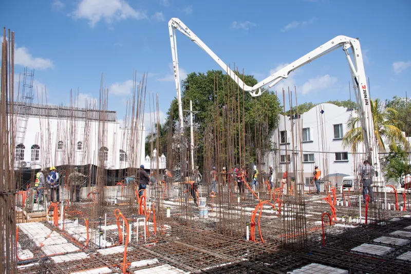An elevated view of the Abund construction site in Riviera Maya, showing a large white 'Parma' concrete pump truck with its boom extended, actively pouring concrete into a vast, newly formed structural slab. Numerous construction workers in safety...