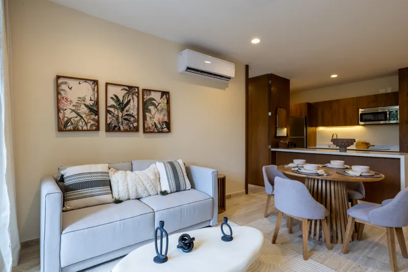 An interior view of Constelada's 1-bedroom model unit, showcasing a modern living and dining area. The space features a light grey sofa with decorative pillows, a sculptural white coffee table, and a round wooden dining table with four light grey ...