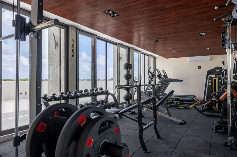 A fully completed and furnished rooftop amenity area at Constelada, featuring a dark-tiled pool with integrated wooden decking, comfortable light-colored beanbag chairs, and lush tropical plants, all set against a vibrant blue sky with white cloud...