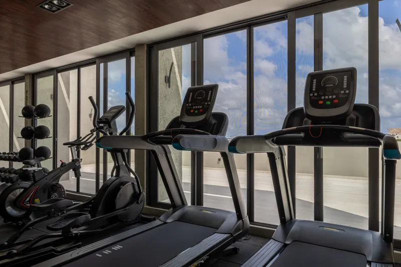 A fully completed and furnished rooftop amenity area at Constelada, featuring a dark-tiled pool with integrated wooden decking, comfortable light-colored beanbag chairs, and lush tropical plants, all set against a vibrant blue sky with white cloud...