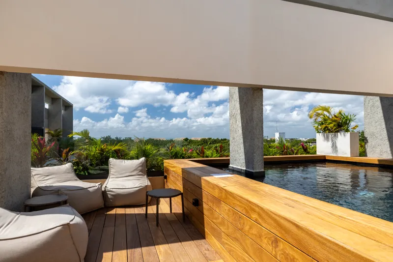 A fully completed and furnished rooftop amenity area at Constelada, featuring a dark-tiled pool with integrated wooden decking, comfortable light-colored beanbag chairs, and lush tropical plants, all set against a vibrant blue sky with white cloud...