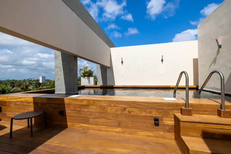 A fully completed and furnished rooftop amenity area at Constelada, featuring a dark-tiled pool with integrated wooden decking, comfortable light-colored beanbag chairs, and lush tropical plants, all set against a vibrant blue sky with white cloud...