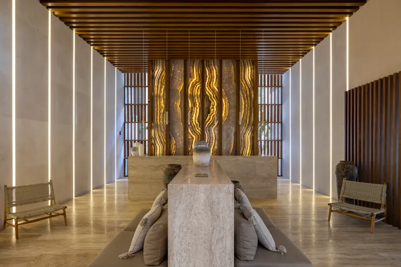 A fully completed and furnished rooftop amenity area at Constelada, featuring a dark-tiled pool with integrated wooden decking, comfortable light-colored beanbag chairs, and lush tropical plants, all set against a vibrant blue sky with white cloud...