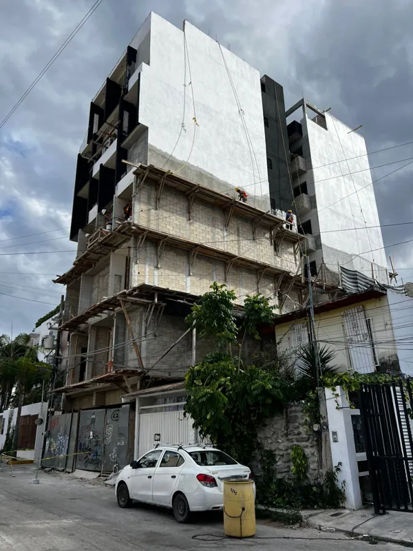 Low-angle view of the B Savage development building under construction in Playa del Carmen. The multi-story building shows its main concrete structure complete, with white exterior plaster applied to the upper floors and dark facade elements visib...