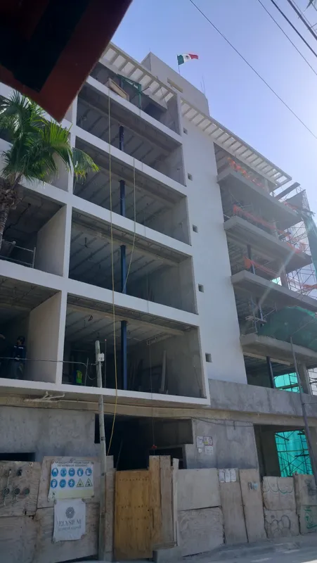 A low-angle view of the Elysium at Fifth Avenue construction site in Playa del Carmen, showing a multi-story building with its concrete structure largely complete and partially covered in white exterior plaster. A tall palm tree stands in front, a...