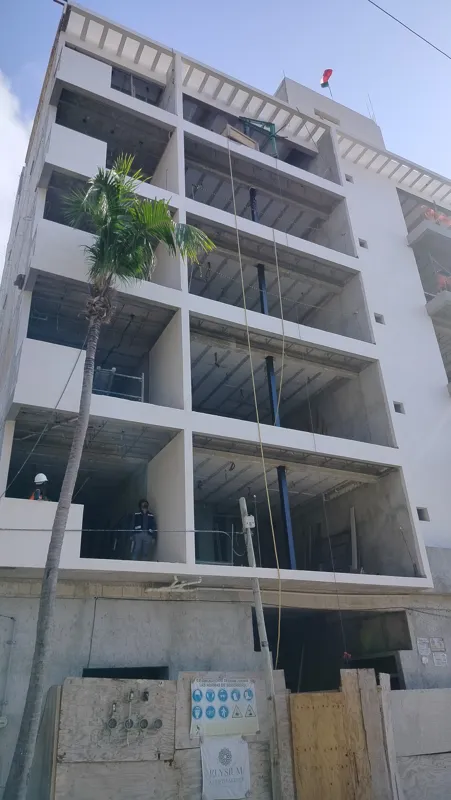 A low-angle view of the Elysium at Fifth Avenue construction site in Playa del Carmen, showing a multi-story building with its concrete structure largely complete and partially covered in white exterior plaster. A tall palm tree stands in front, a...