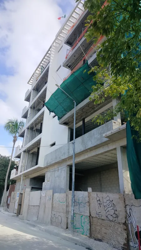A low-angle view of the Elysium at Fifth Avenue construction site in Playa del Carmen, showing a multi-story building with its concrete structure largely complete and partially covered in white exterior plaster. A tall palm tree stands in front, a...