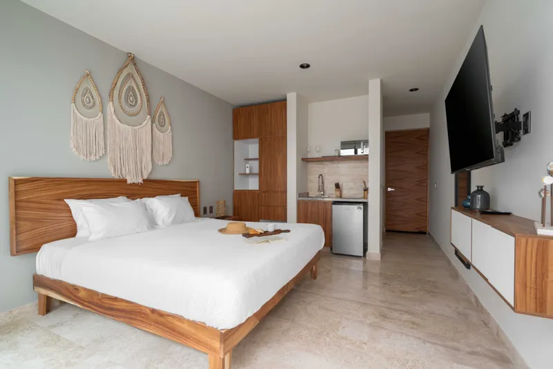 A beautifully staged studio model unit at Rio Tulum, featuring a queen-sized bed with white linens, a built-in wooden wardrobe, a compact kitchenette with a microwave and mini-fridge, a wall-mounted television, and polished light-colored stone flo...