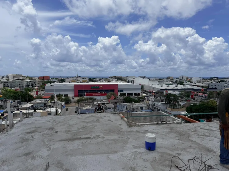 Panoramic view from the newly completed upper-level concrete slab at Ola de Mar, showcasing the urban landscape of Playa del Carmen, including a large commercial building, and the distant Caribbean Sea under a blue sky with white clouds. Construct...