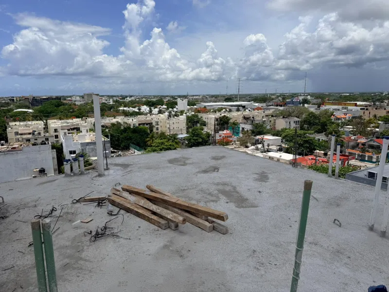 Panoramic view from the newly completed upper-level concrete slab at Ola de Mar, showcasing the urban landscape of Playa del Carmen, including a large commercial building, and the distant Caribbean Sea under a blue sky with white clouds. Construct...