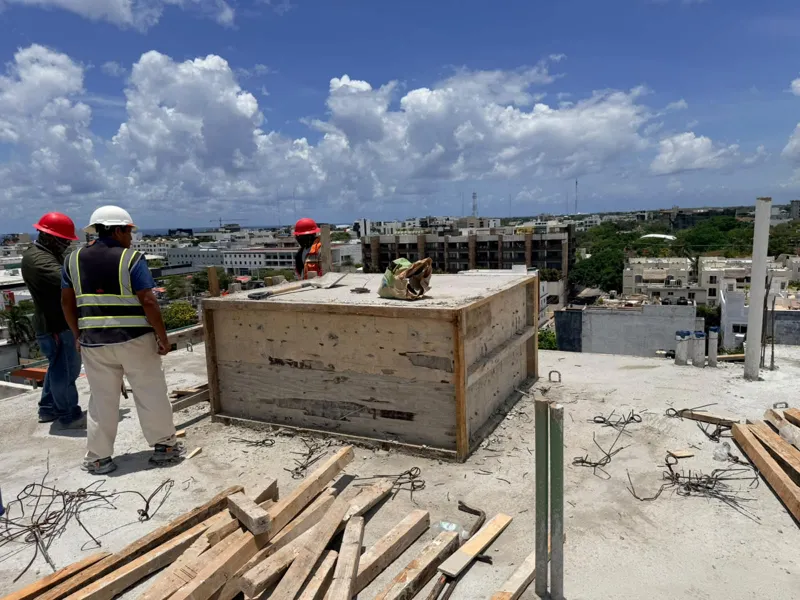 Panoramic view from the newly completed upper-level concrete slab at Ola de Mar, showcasing the urban landscape of Playa del Carmen, including a large commercial building, and the distant Caribbean Sea under a blue sky with white clouds. Construct...