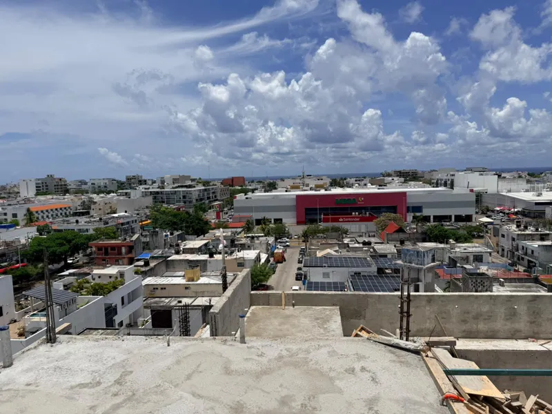 Panoramic view from the newly completed upper-level concrete slab at Ola de Mar, showcasing the urban landscape of Playa del Carmen, including a large commercial building, and the distant Caribbean Sea under a blue sky with white clouds. Construct...