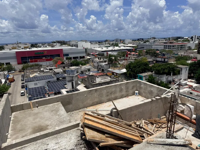 Panoramic view from the newly completed upper-level concrete slab at Ola de Mar, showcasing the urban landscape of Playa del Carmen, including a large commercial building, and the distant Caribbean Sea under a blue sky with white clouds. Construct...