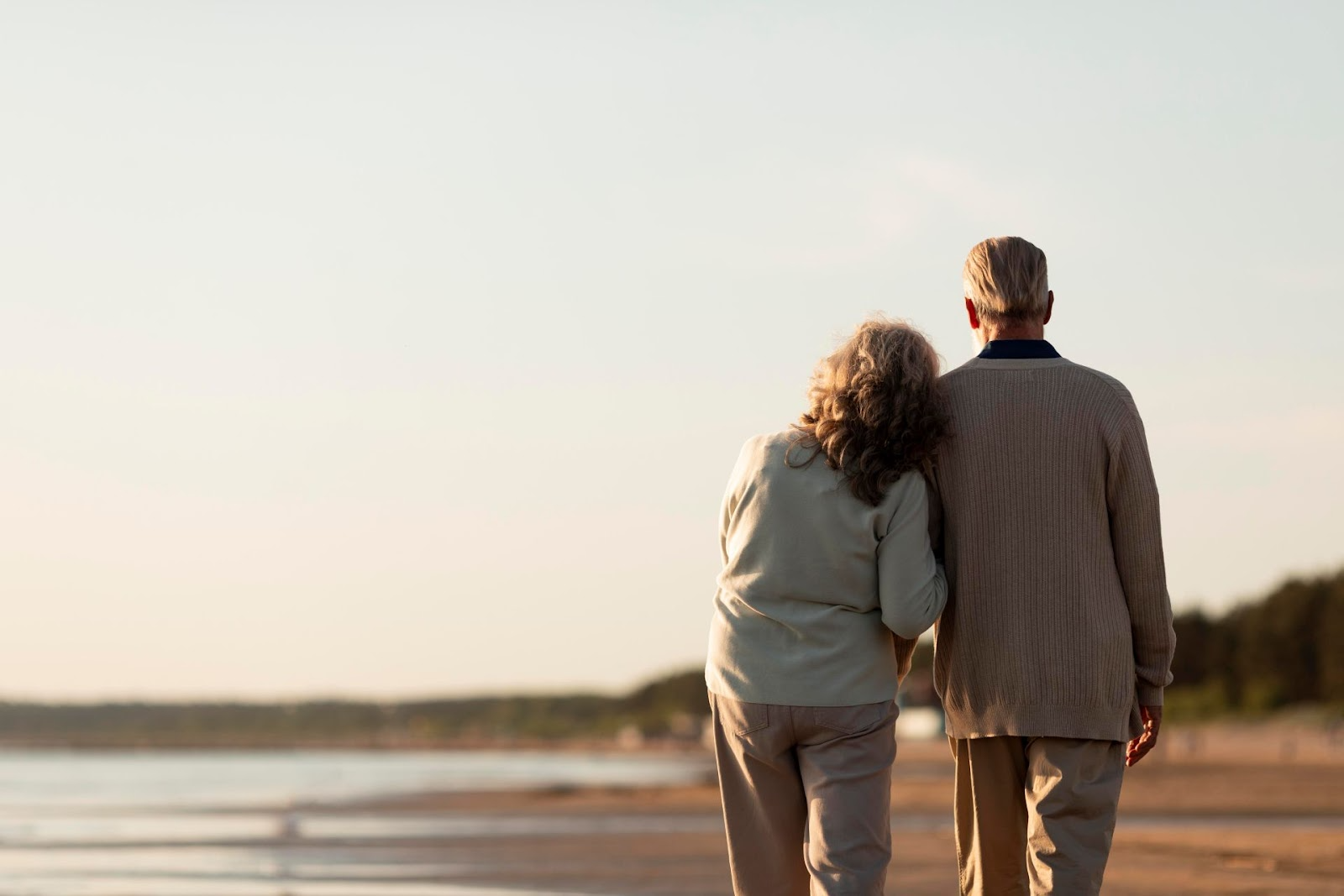 Pareja madura caminando abrazada en la playa al atardecer, representando jubilación feliz