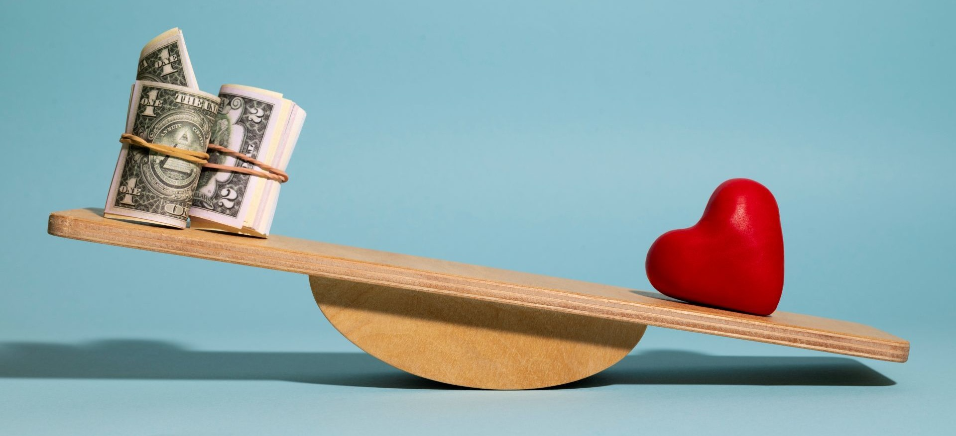 Balanza de madera equilibrando dinero enrollado y corazón rojo en fondo azul