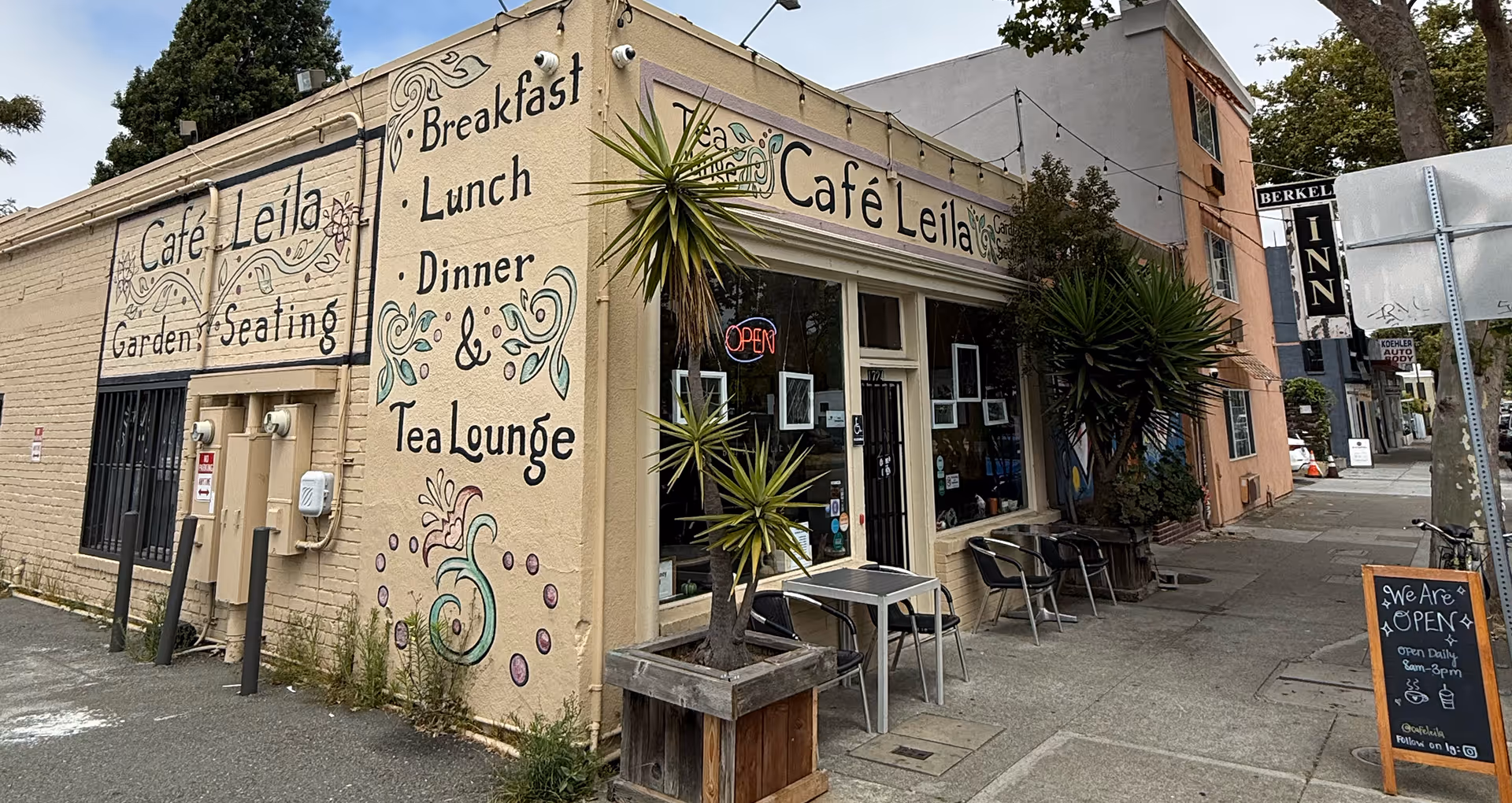Corner view of Café Leila building with garden seating, outdoor tables, palm plants, and signs indicating breakfast, lunch, dinner, and tea lounge.