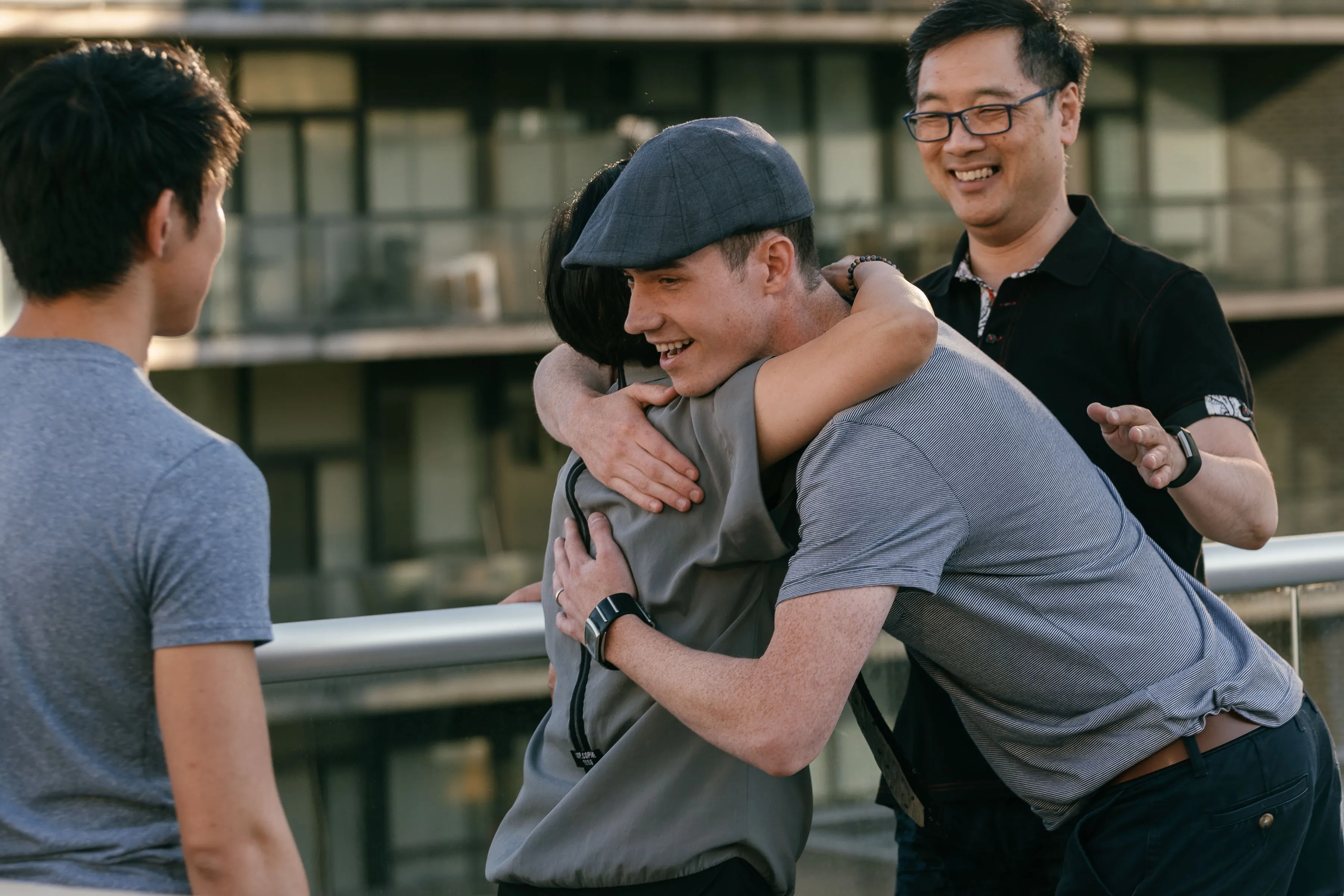 Two people embracing outdoors while another man watches with a smile. A fourth person stands nearby, engaged in the interaction. The scene takes place on a balcony with modern buildings in the background.