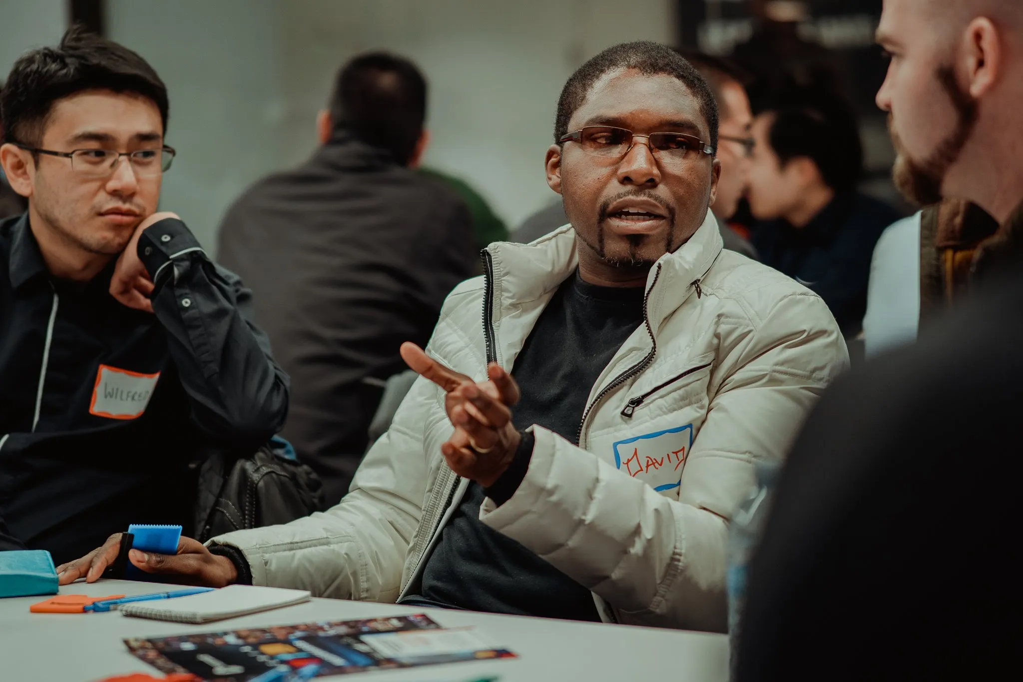 A group of people engaged in a discussion at a round table. A man wearing a white jacket gestures while speaking, and another man in black glasses listens attentively. Name tags are visible on their clothing.