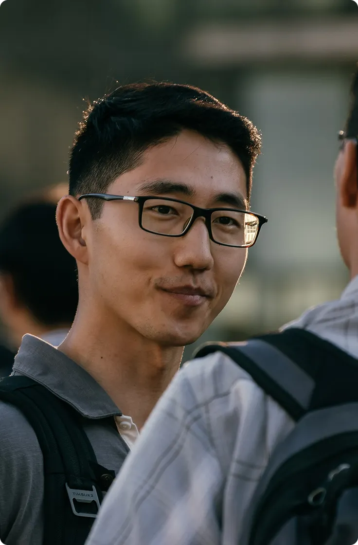 A young man with glasses and a short hairstyle, wearing a gray polo shirt, is smiling slightly while looking at another person in a casual outdoor setting. The background is blurred, suggesting a social environment.