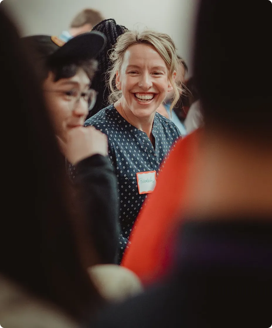 A group of people engaged in conversation, with a woman in the foreground smiling. She wears a blue patterned shirt and a name tag. The background is slightly blurred, capturing a lively atmosphere.