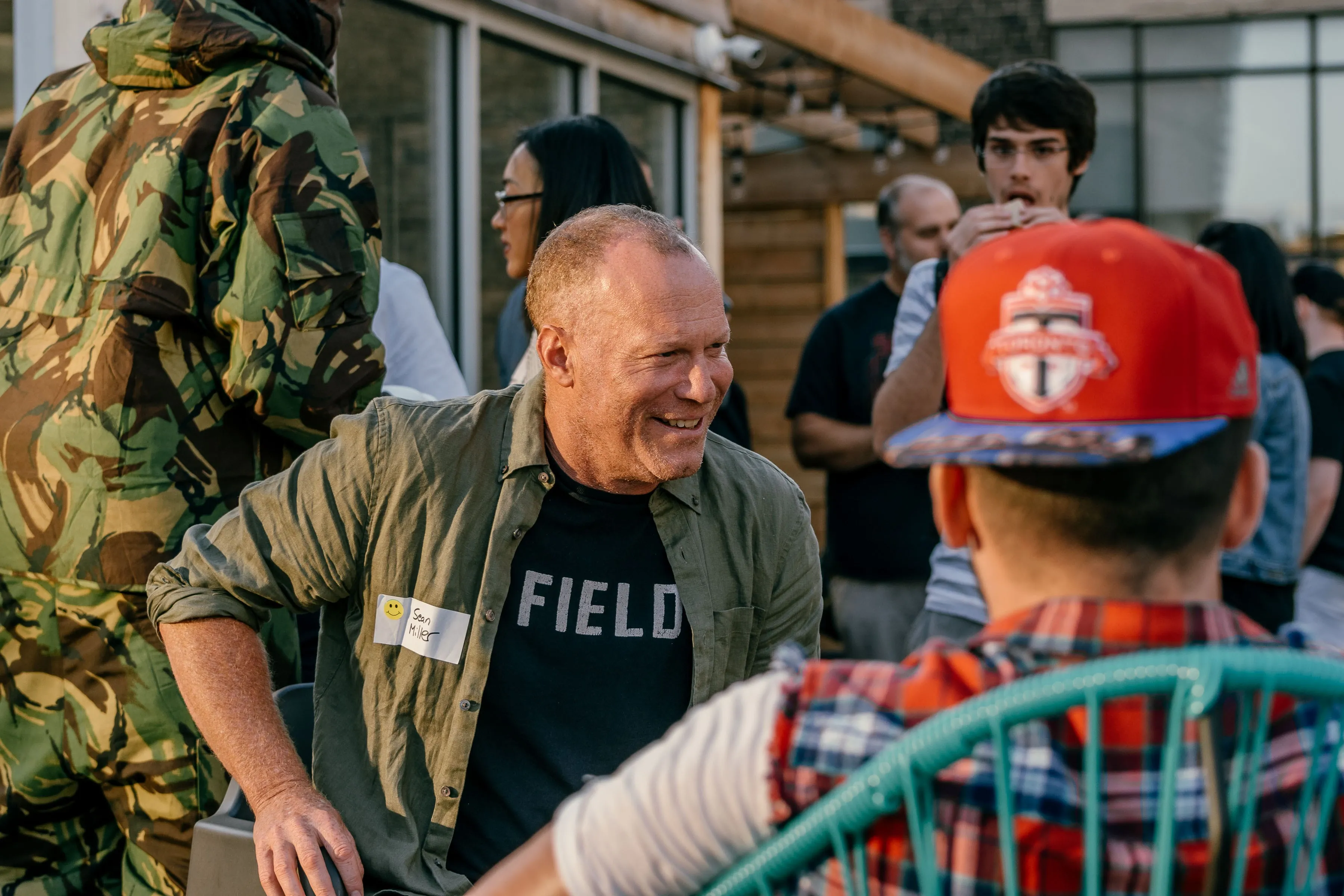 A man in a green shirt and glasses smiling while talking with another man wearing a red cap. Several people are gathered in the background, enjoying a casual outdoor event.