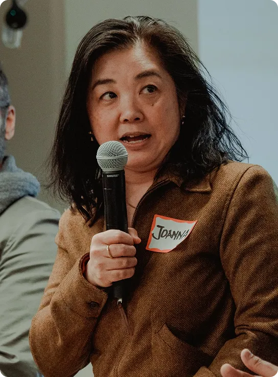 A woman speaking into a microphone during a panel discussion, wearing a brown jacket with a name tag that reads 'Joanna'. The background shows a blurred image of an audience.