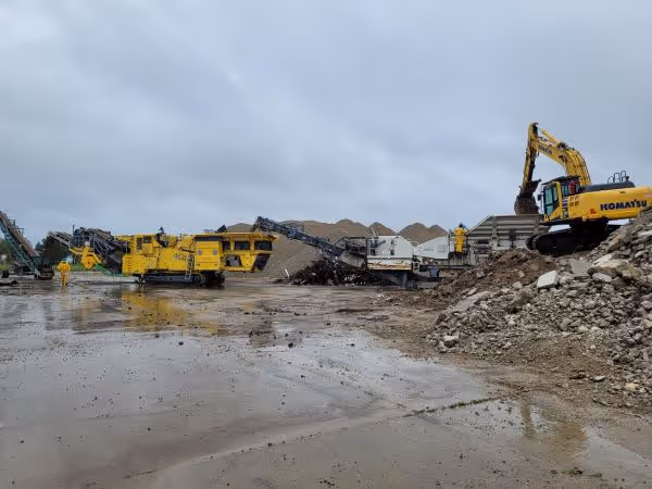 Construction site with yellow Komatsu excavator loading rubble into a crusher machine under an overcast sky.