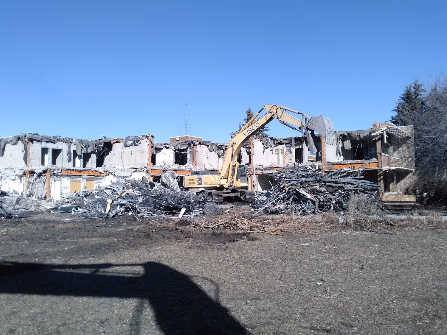 Excavator demolishing a two-story building with piles of rubble in front under a clear blue sky.